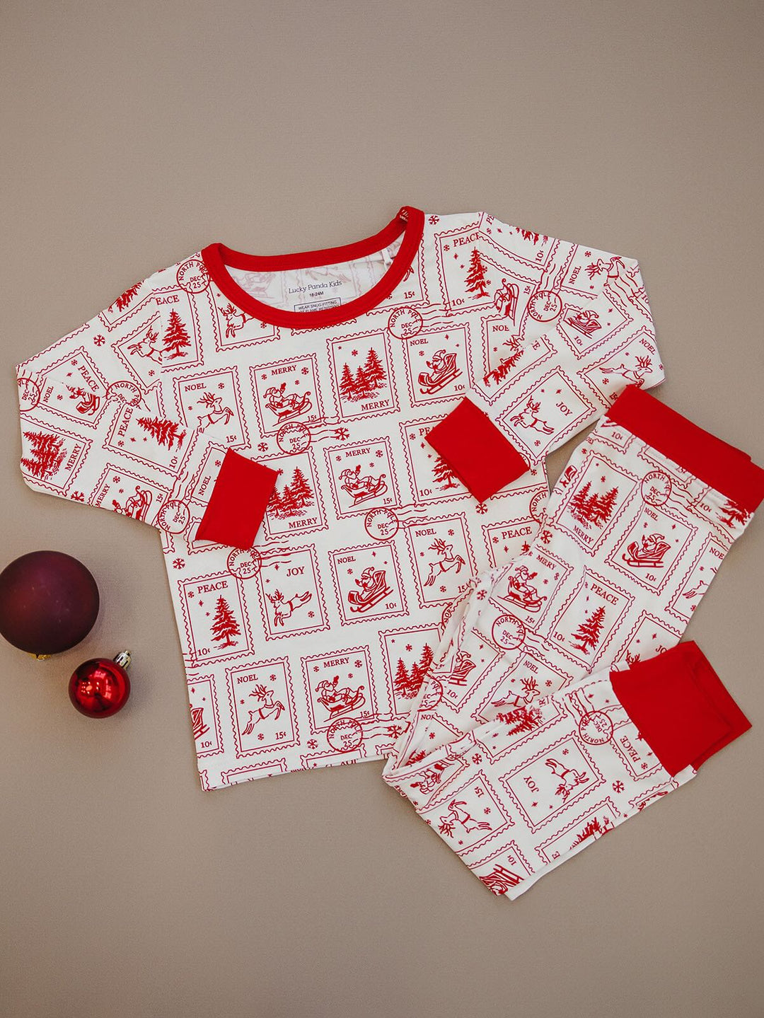 North Pole Stamps two piece Christmas pajamas laid flat beside holiday ornaments, showing red trim and stamp print.