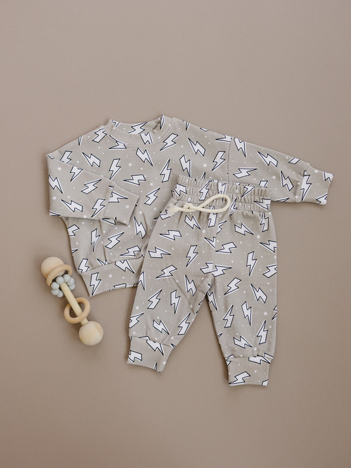 Latte Bolts crewneck top and matching jogger pants laid flat beside a wooden toy, showing the lightning bolt print.