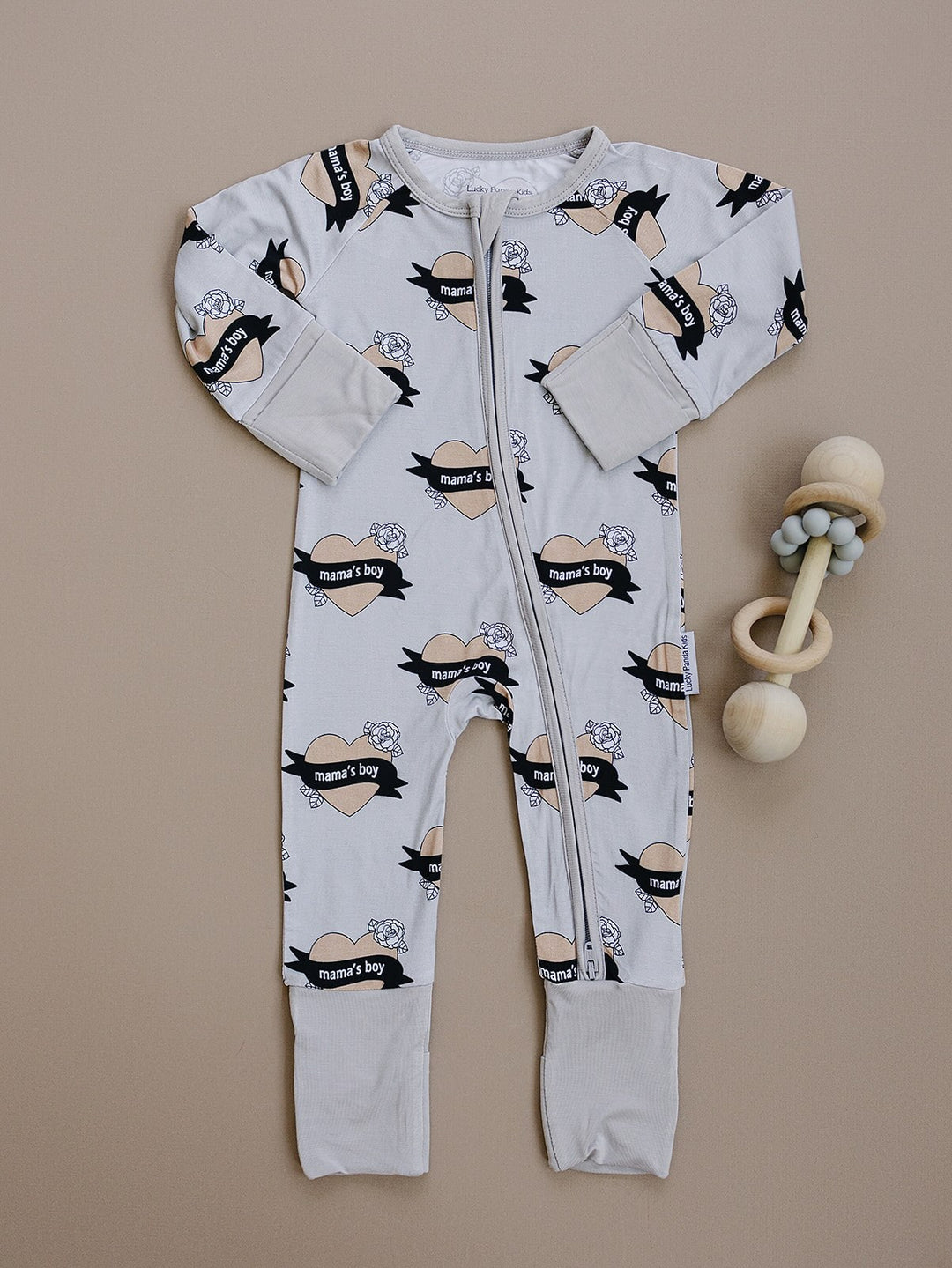 Mama’s Boy long-sleeve bamboo zip romper with beige hearts and black banners displayed flat.