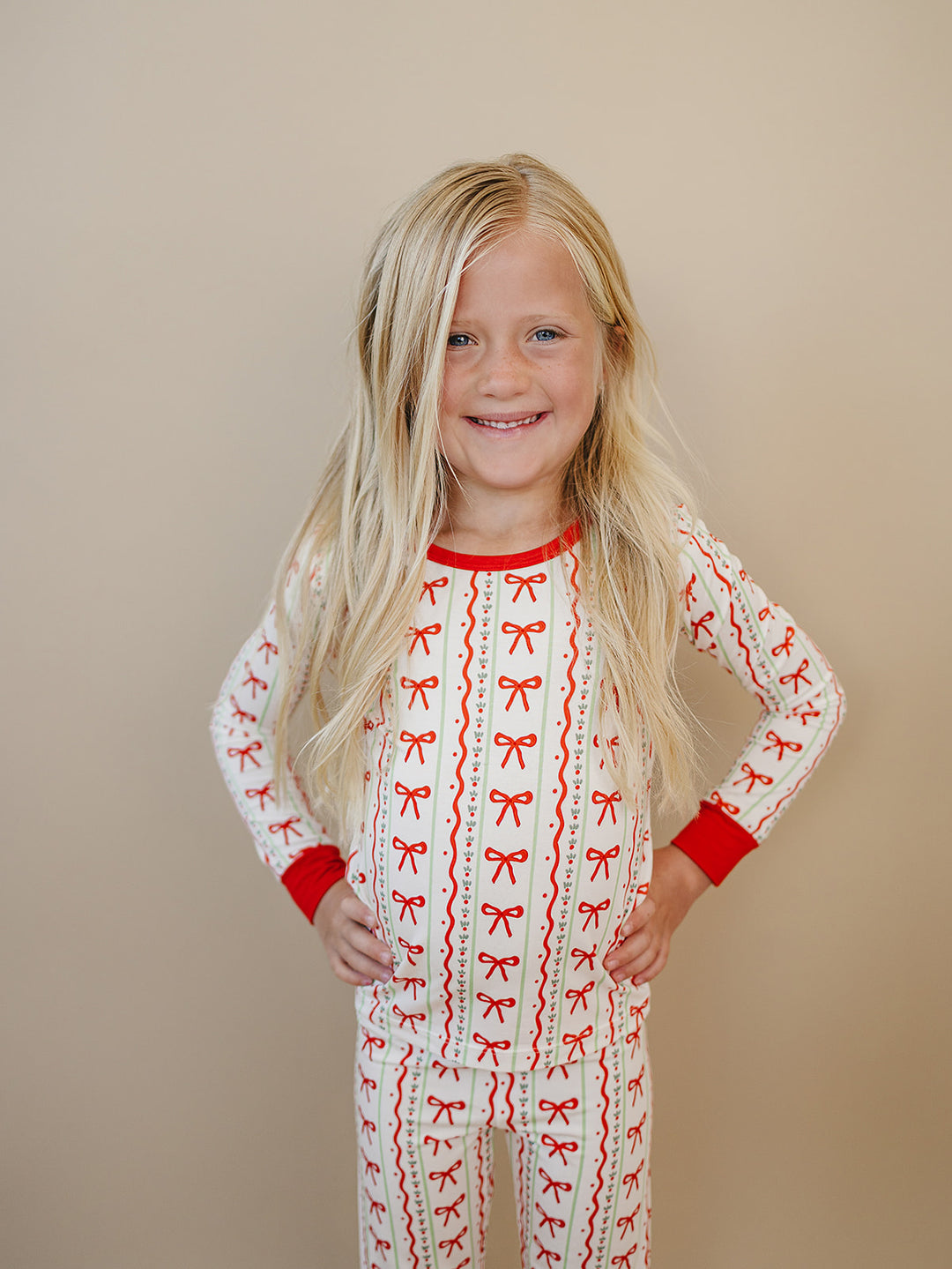 Smiling child with hands on hips wearing Christmas Bows pajamas with red bows, decorative stripes and red trim.