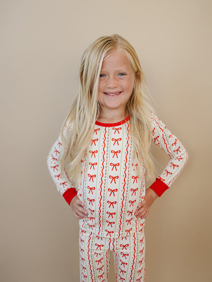 Smiling child with hands on hips wearing Christmas Bows pajamas with red bows, decorative stripes and red trim.