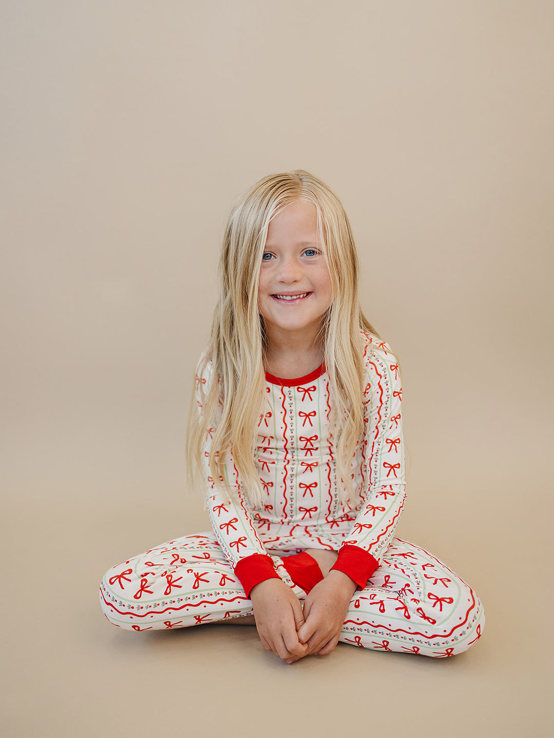 Smiling child sitting on the floor with legs crossed while wearing Christmas Bows two piece pajamas.