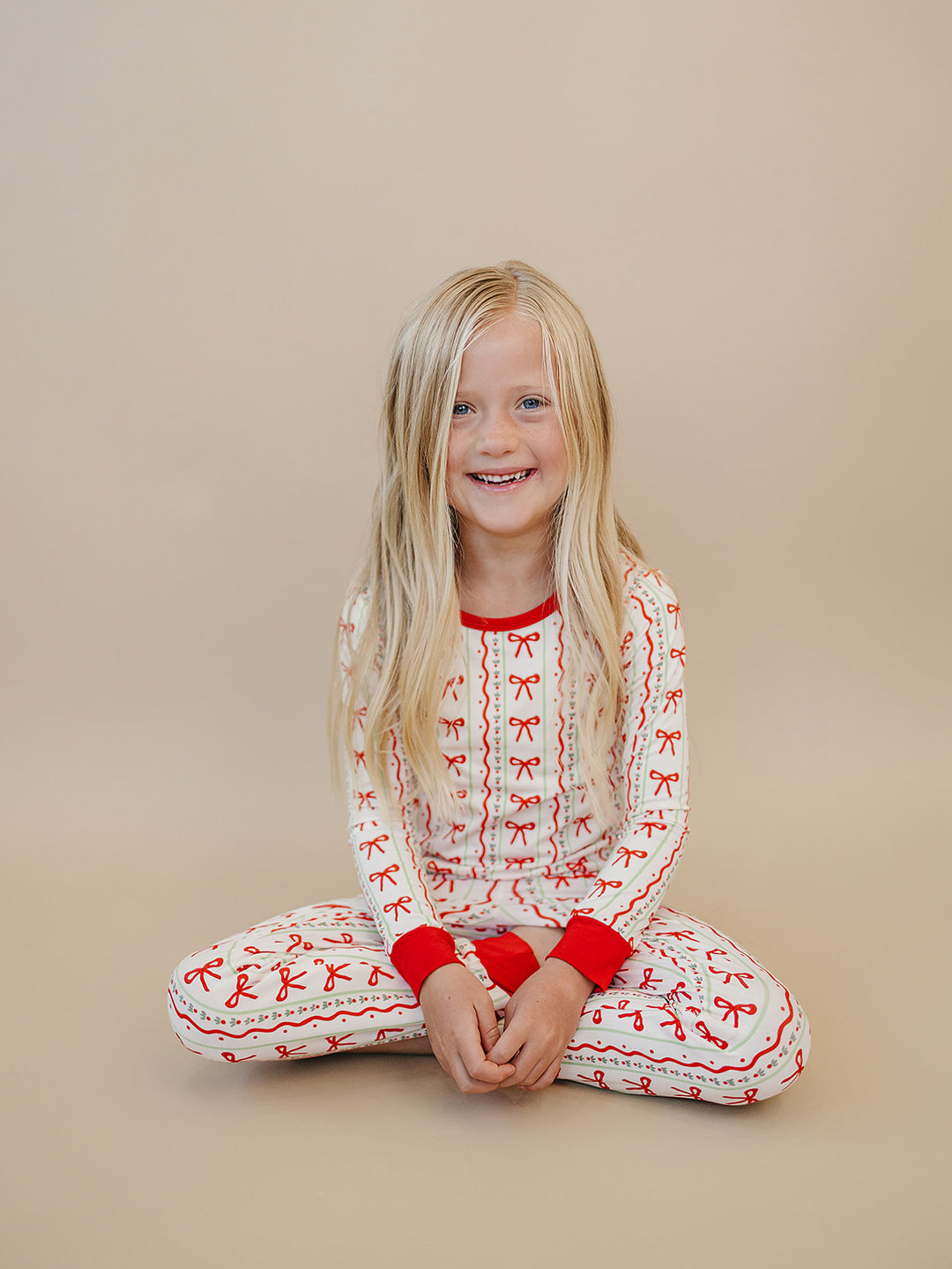 Child sitting with legs crossed in the Christmas Bows pajama top and matching pants with red cuffs.