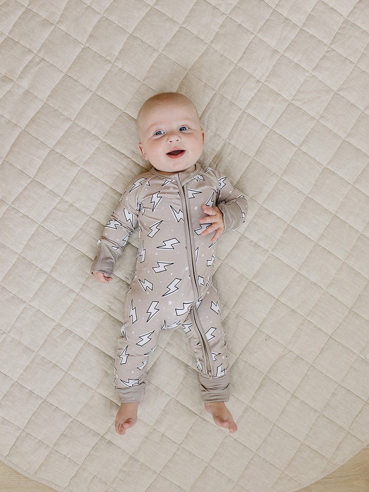 Smiling baby lying on a cream quilt in the Latte Bolts bamboo zip romper in a full-body view.