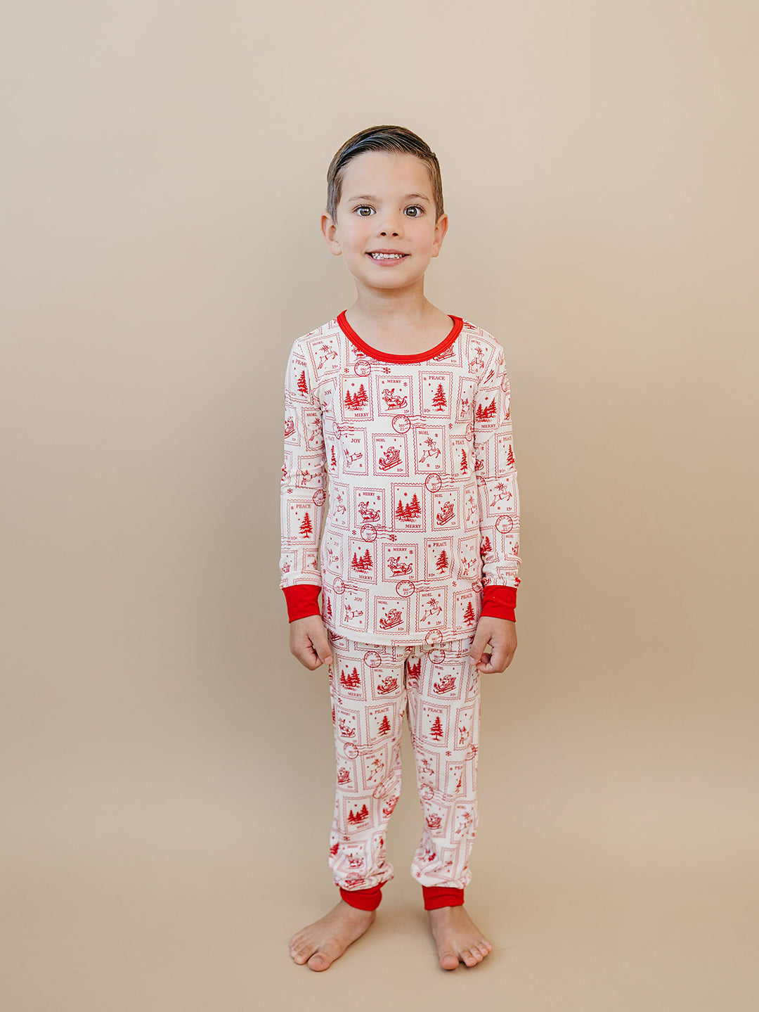 Child smiling in North Pole Stamps two piece pajamas with a cream background and red holiday print.