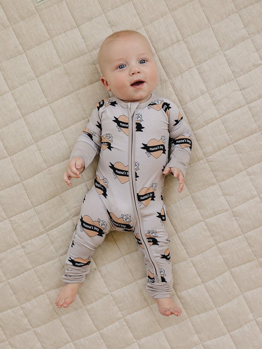 Smiling baby lying on a cream quilt in the Mama’s Boy bamboo zip romper with beige hearts and black banners.