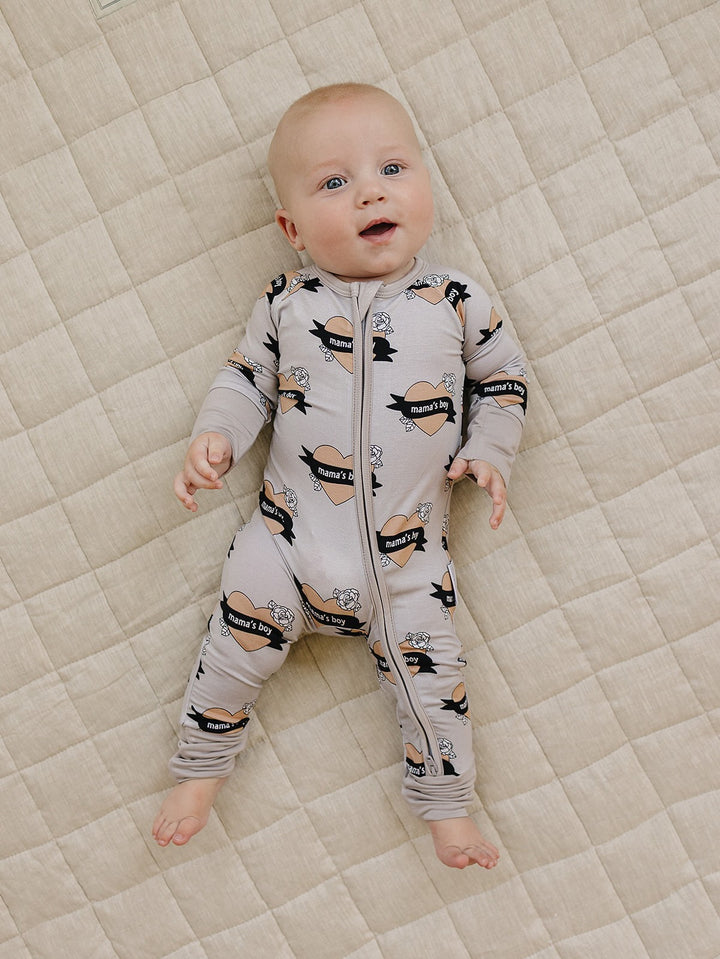 Smiling baby lying on a cream quilt in the Mama’s Boy bamboo zip romper with beige hearts and black banners.