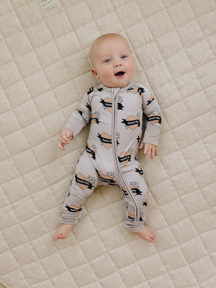 Full-length view of a baby wearing the Mama’s Boy bamboo zip romper on a cream quilt.