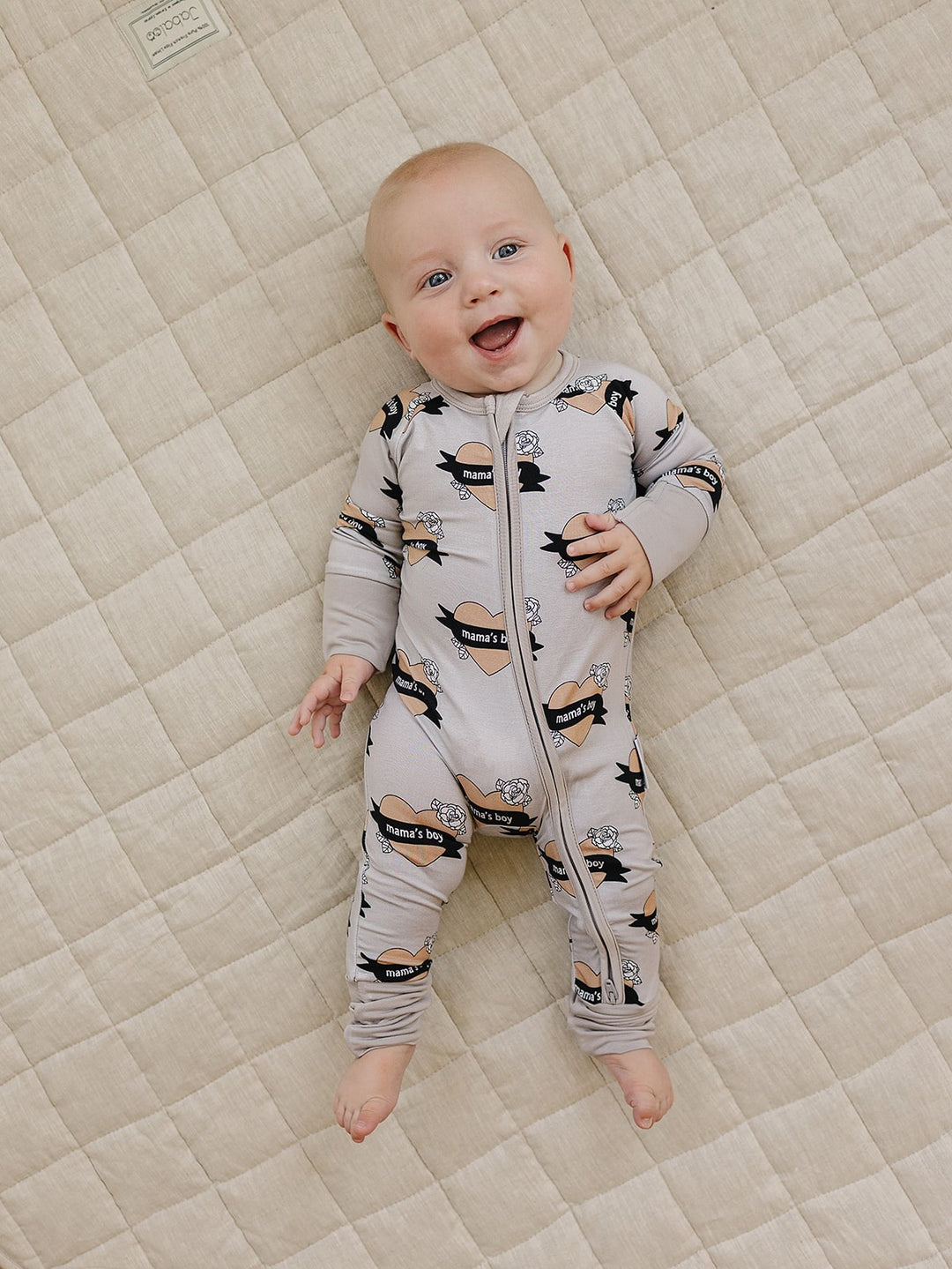 Laughing baby lying on a cream quilt in the Mama’s Boy bamboo zip romper.