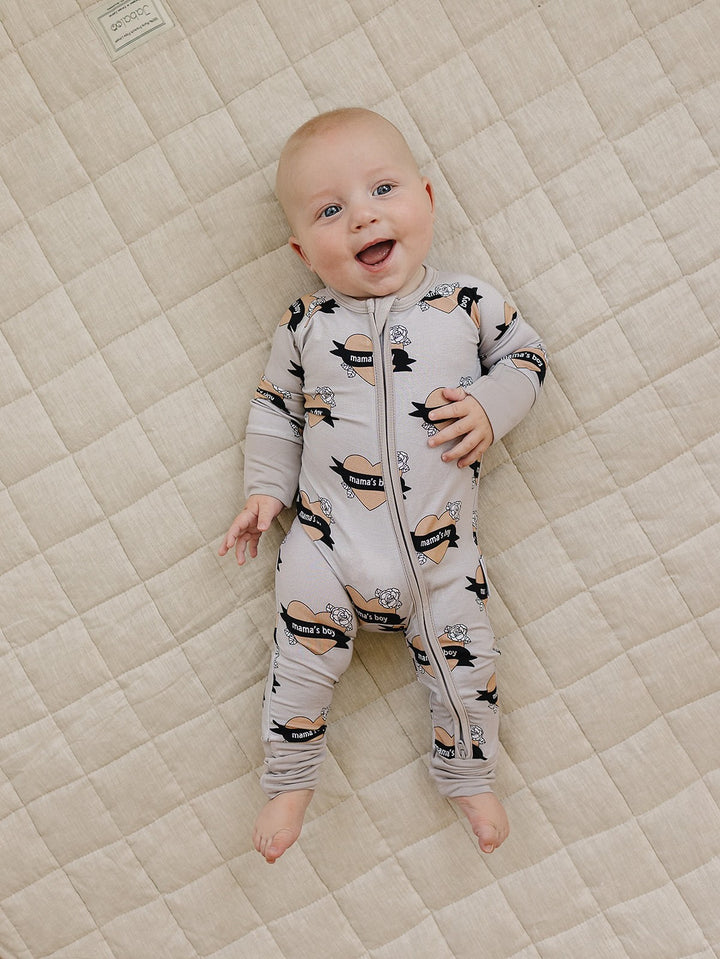 Laughing baby lying on a cream quilt in the Mama’s Boy bamboo zip romper.