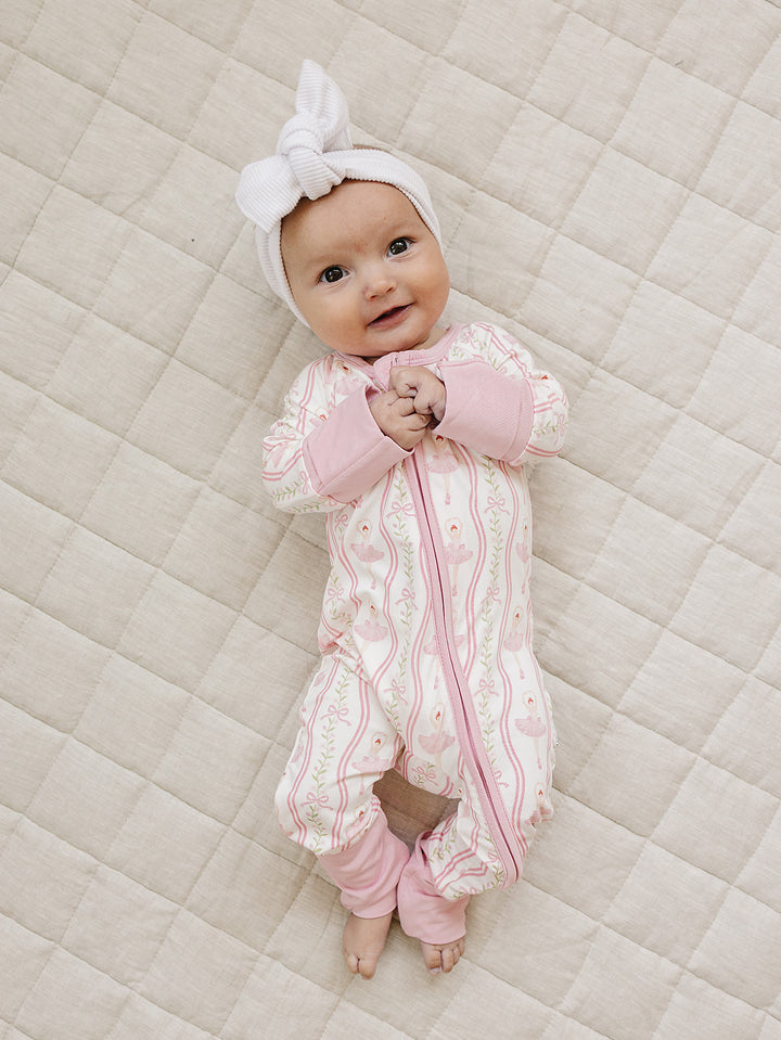 Smiling baby wearing the Little Ballerina bamboo zip romper and a white bow headband on a cream quilted mat.