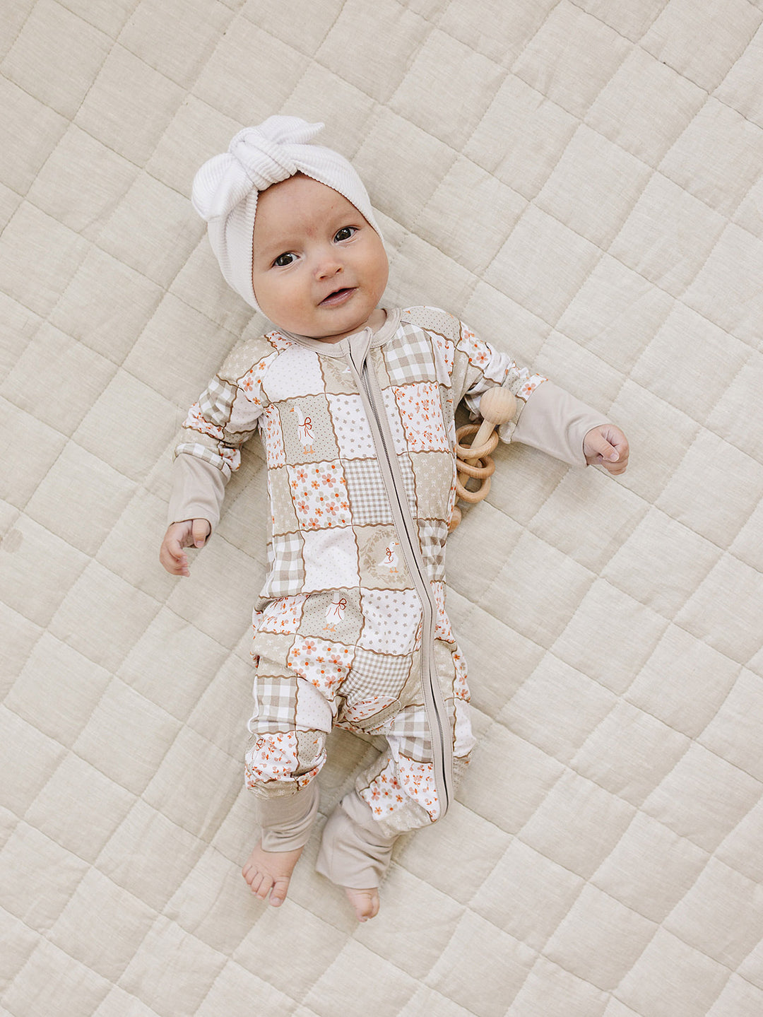 Baby wearing the Lucky Panda Kids Cottage Goose bamboo zip romper and a white bow headwrap while lying on a cream quilt.