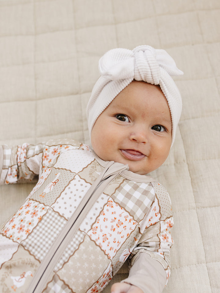 Smiling baby in a white bow headwrap wearing the Cottage Goose bamboo zip romper on a quilted blanket.