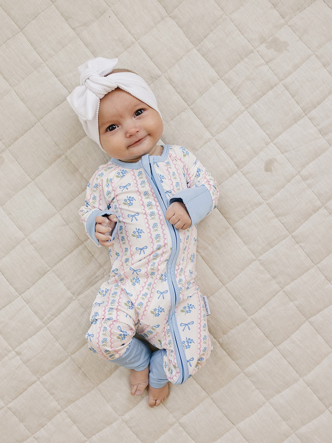 Smiling baby wearing the Whimsy Bloom bamboo zip romper and white bow headband on a cream quilt.