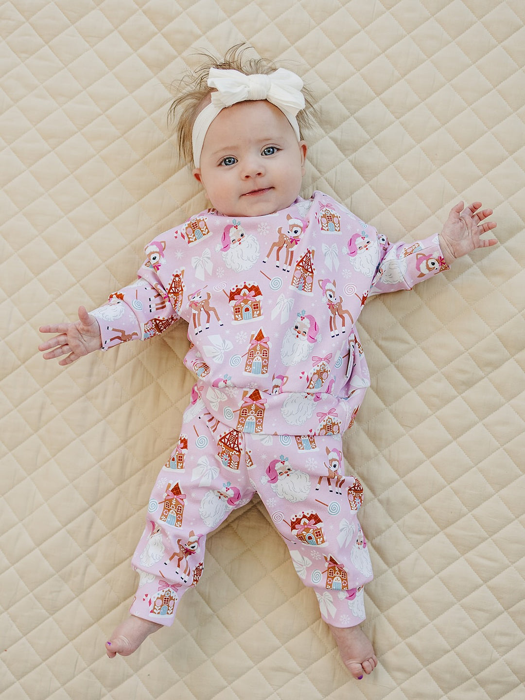 Baby wearing a white bow headband and Pink Christmas jogger set with legs stretched out on a quilt.