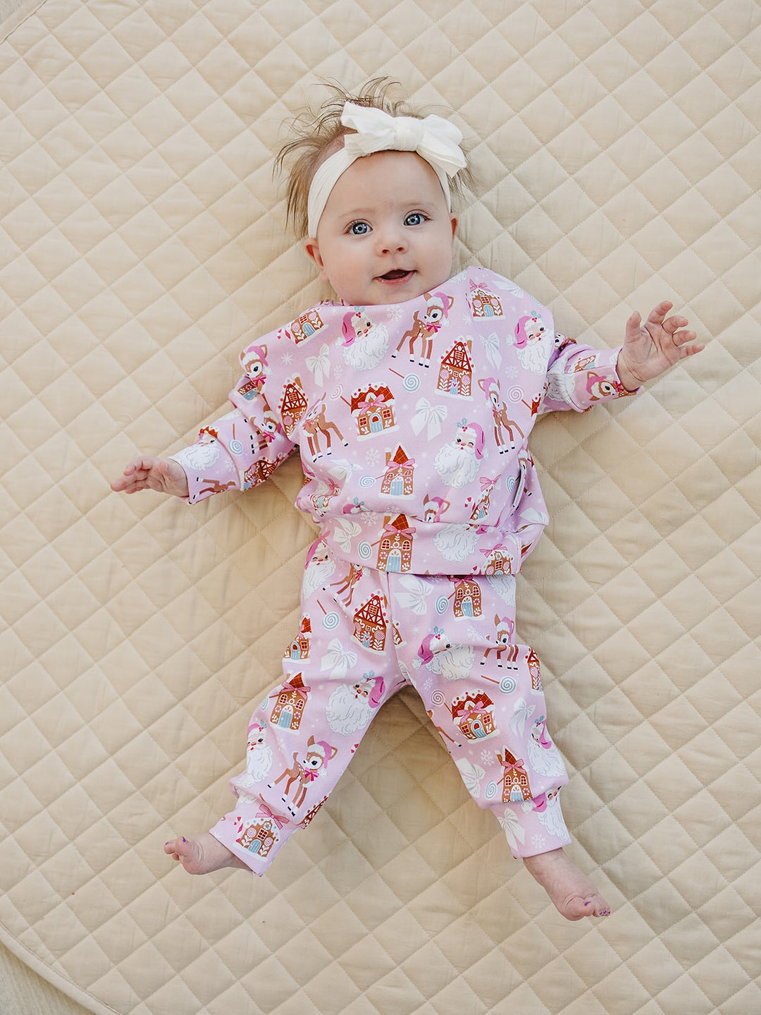Baby wearing the Pink Christmas jogger set and a white bow headband while lying on a cream quilt.