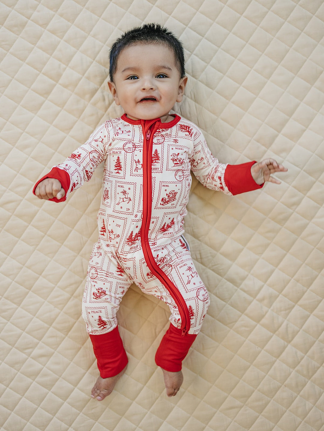 Baby stretching out on a cream quilt in the North Pole Stamps romper with red holiday stamps.