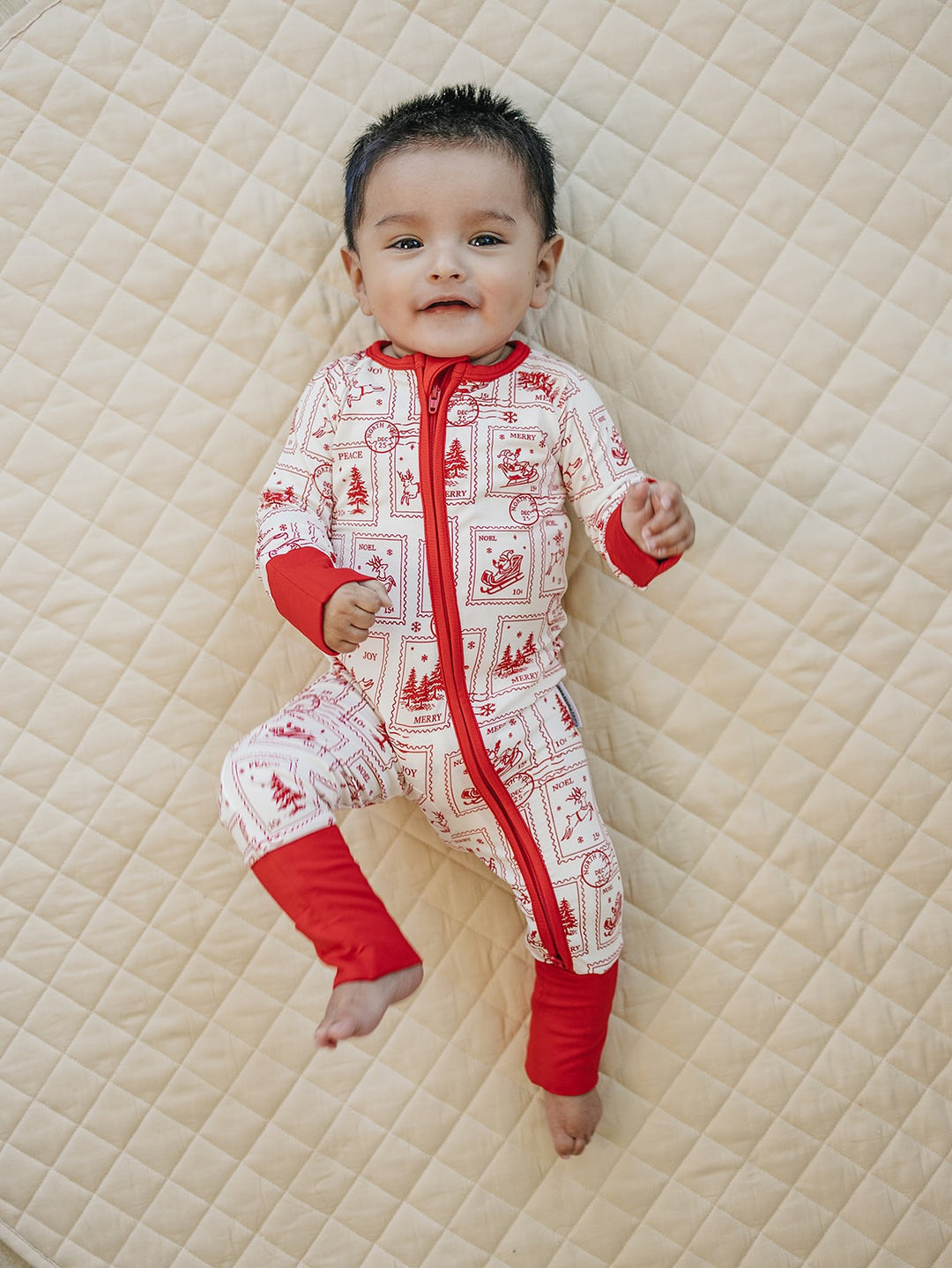 Baby lying on a cream quilt wearing the North Pole Stamps Christmas zip romper with red cuffs and trim.