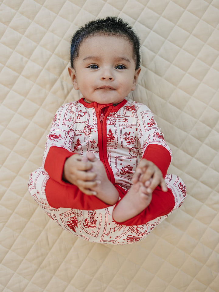 Baby holding their feet while wearing the North Pole Stamps Christmas zip romper on a cream quilt.