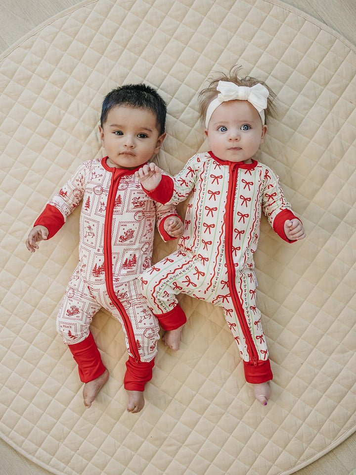 Two babies lying together on a cream quilt in North Pole Stamps and Christmas Bows zip rompers.