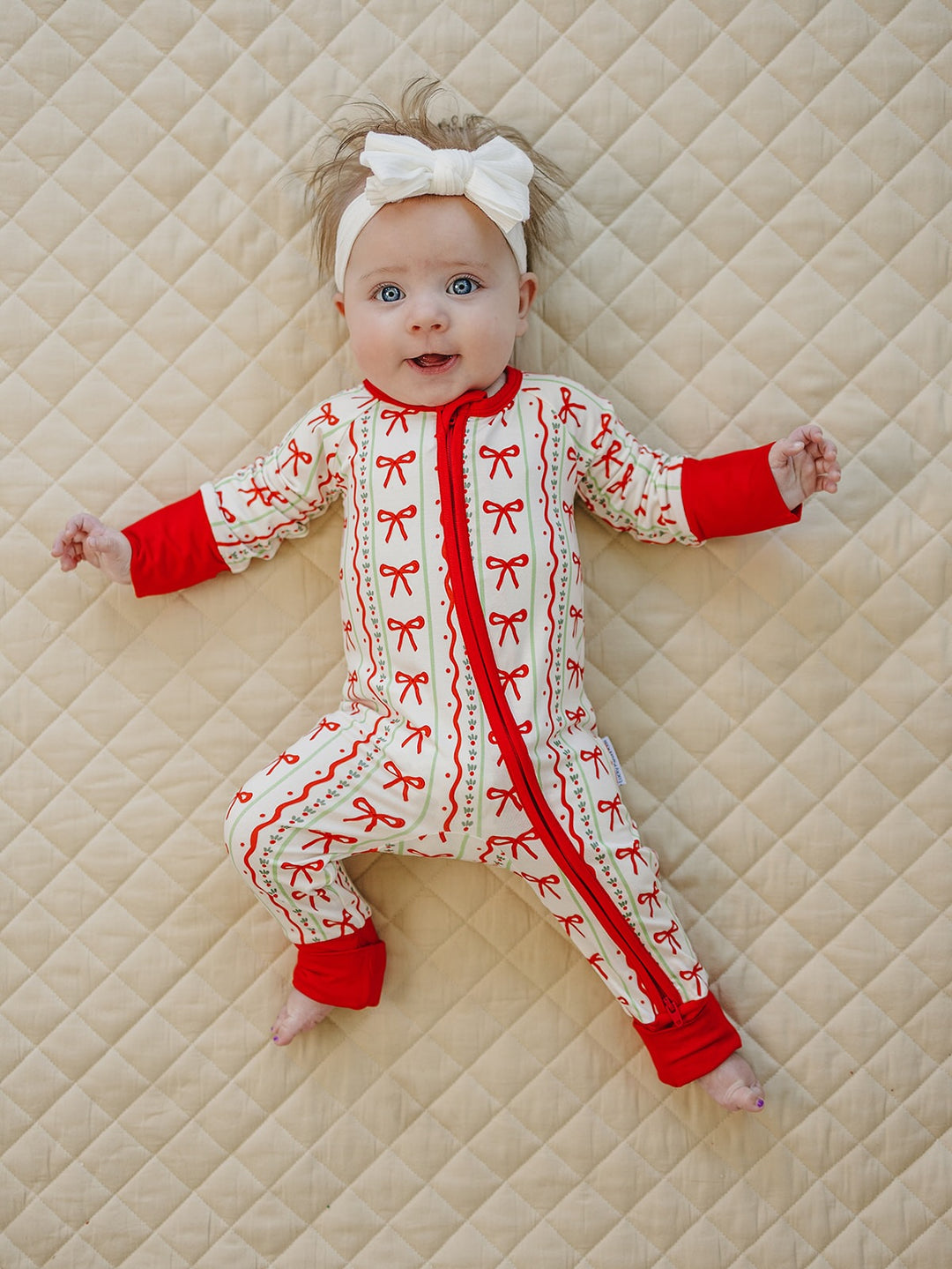 Baby bending one knee on a cream quilt while wearing the Christmas Bows romper and a white bow headband.