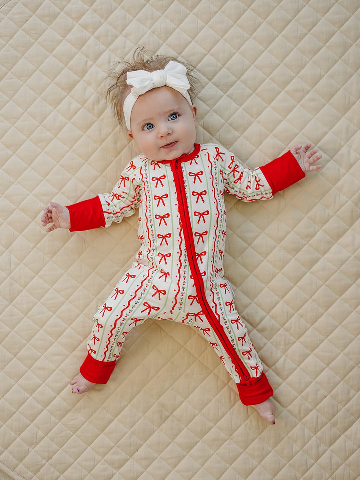 Baby on a cream quilt wearing a white bow headband and Christmas Bows zip romper with red cuffs.