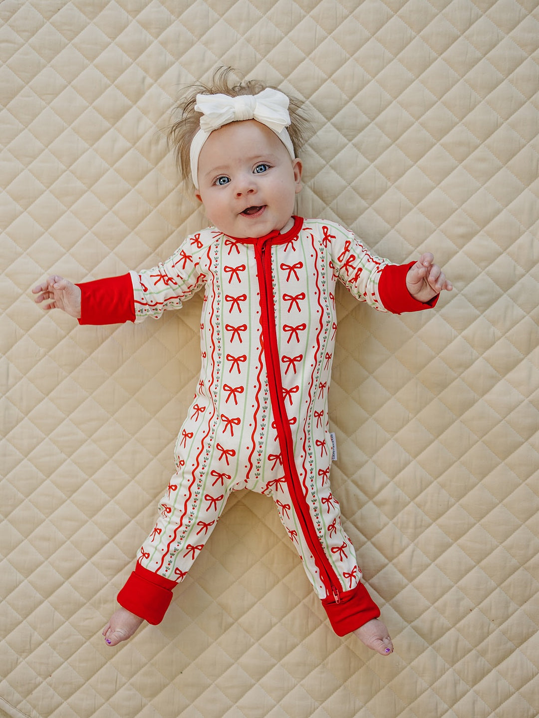 Baby smiling with arms open in the Christmas Bows zip romper with red cuffs on a cream quilt.