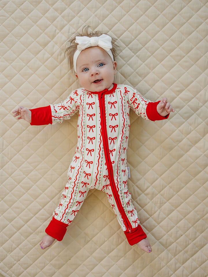 Baby smiling with arms open in the Christmas Bows zip romper with red cuffs on a cream quilt.