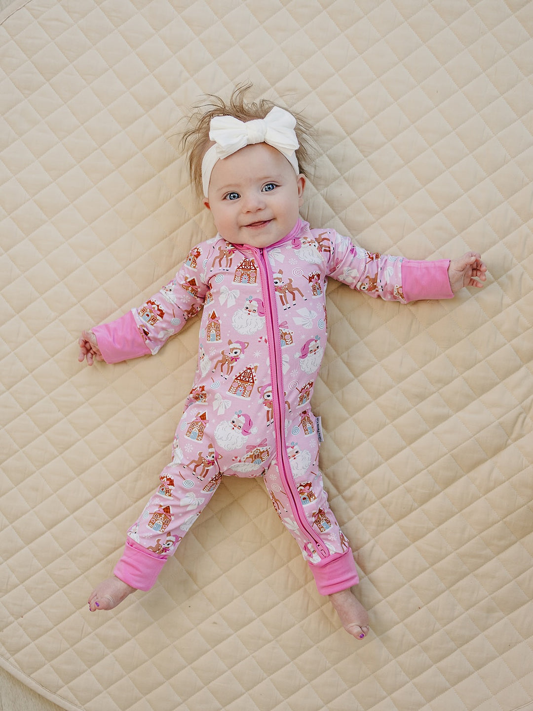 Baby wearing a white bow headband and Pink Christmas zip romper while lying on a cream quilt.