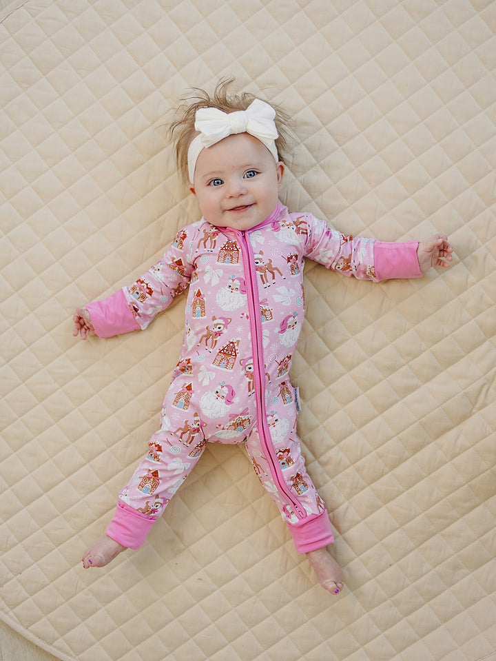 Baby wearing a white bow headband and Pink Christmas zip romper while lying on a cream quilt.