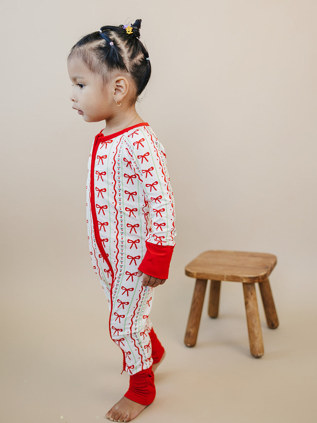Side view of a toddler standing beside a wooden stool in the Christmas Bows zip romper.