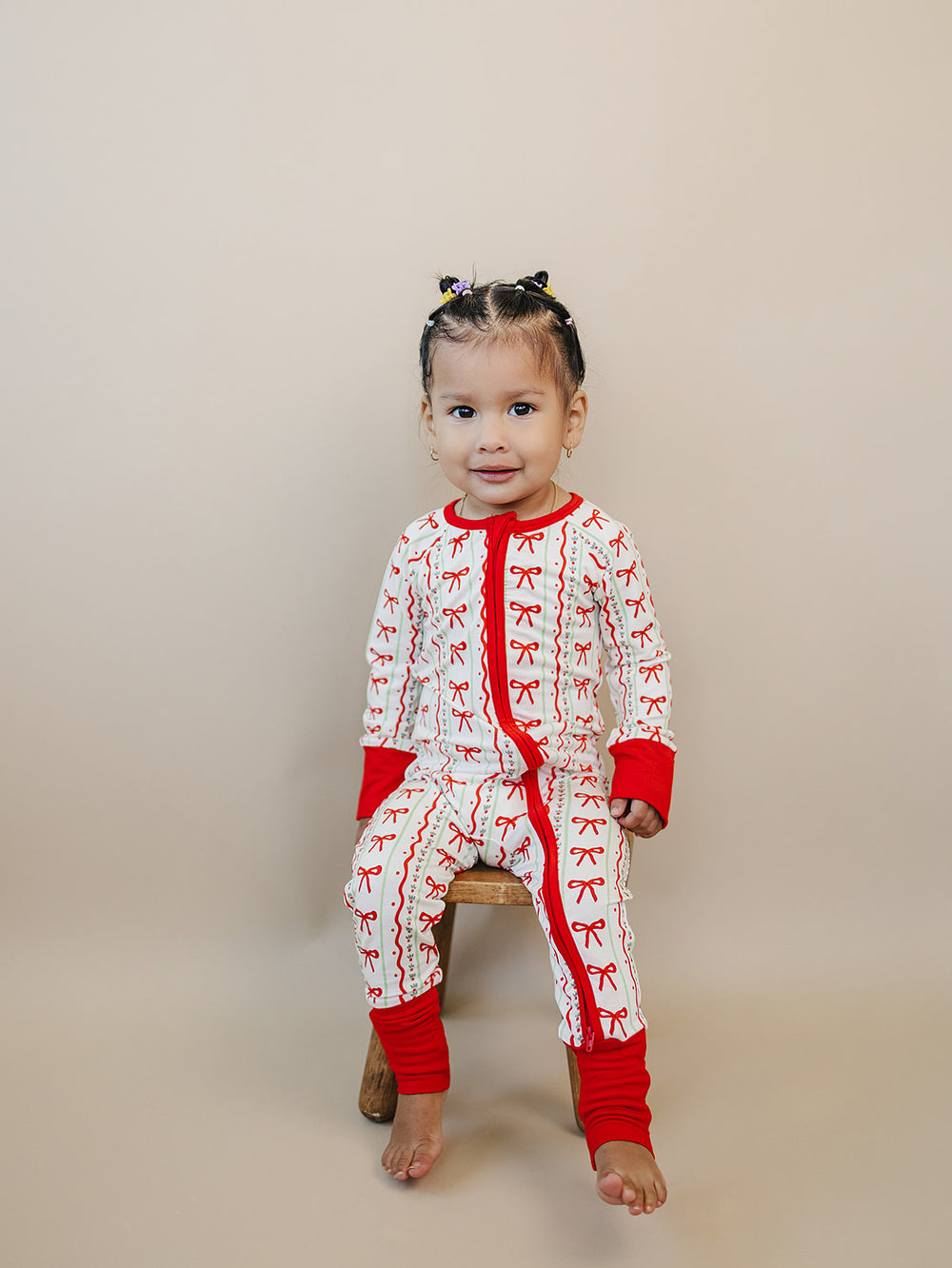 Toddler sitting on a wooden stool in the Christmas Bows zip romper with red wrist and ankle cuffs.
