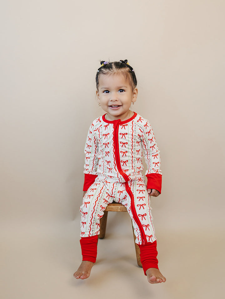 Smiling toddler sitting on a wooden stool in the Christmas Bows romper with bare feet and red ankle cuffs.