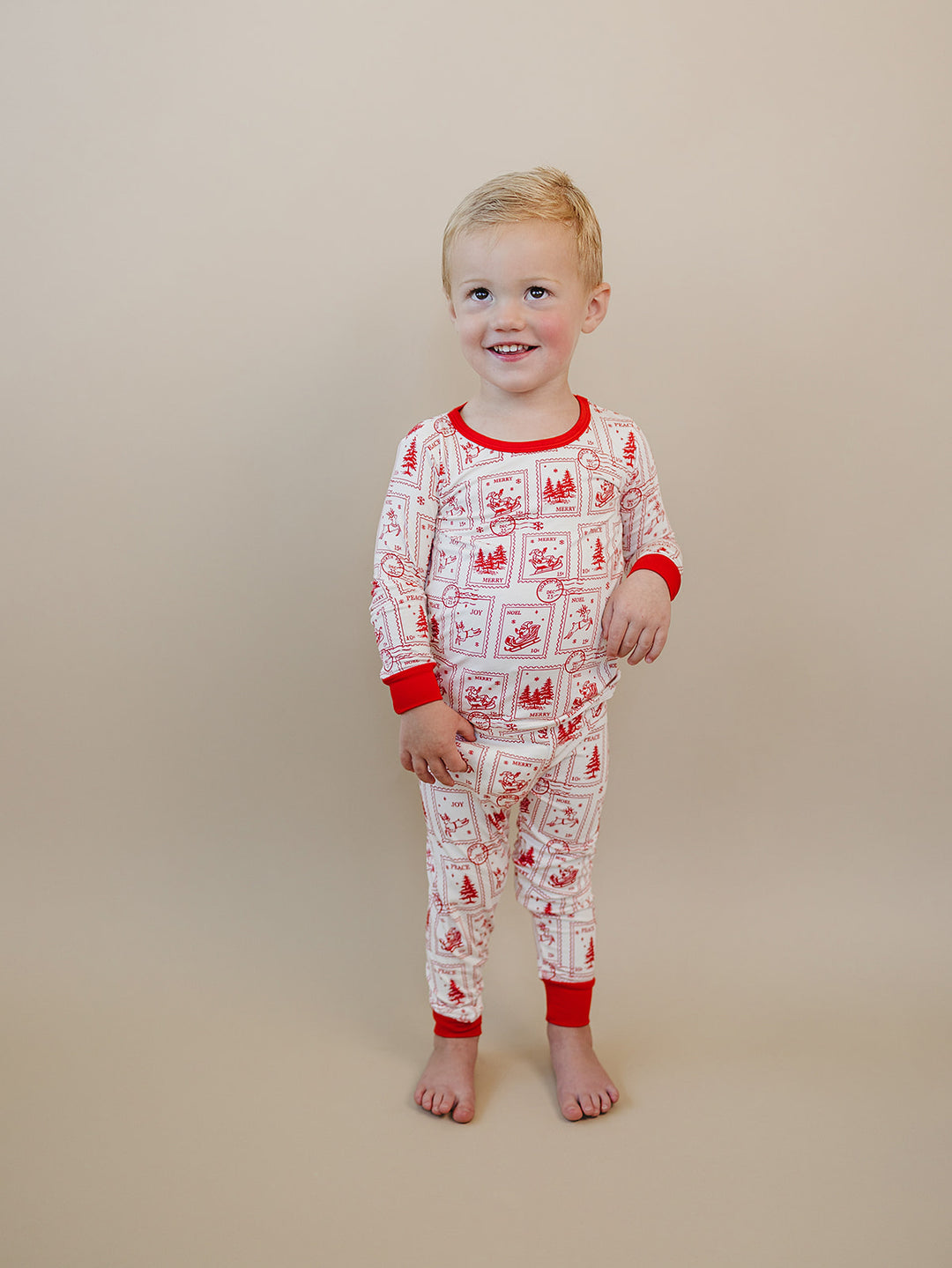 Smiling child wearing cream North Pole Stamps pajamas with red Christmas stamps and matching pants.