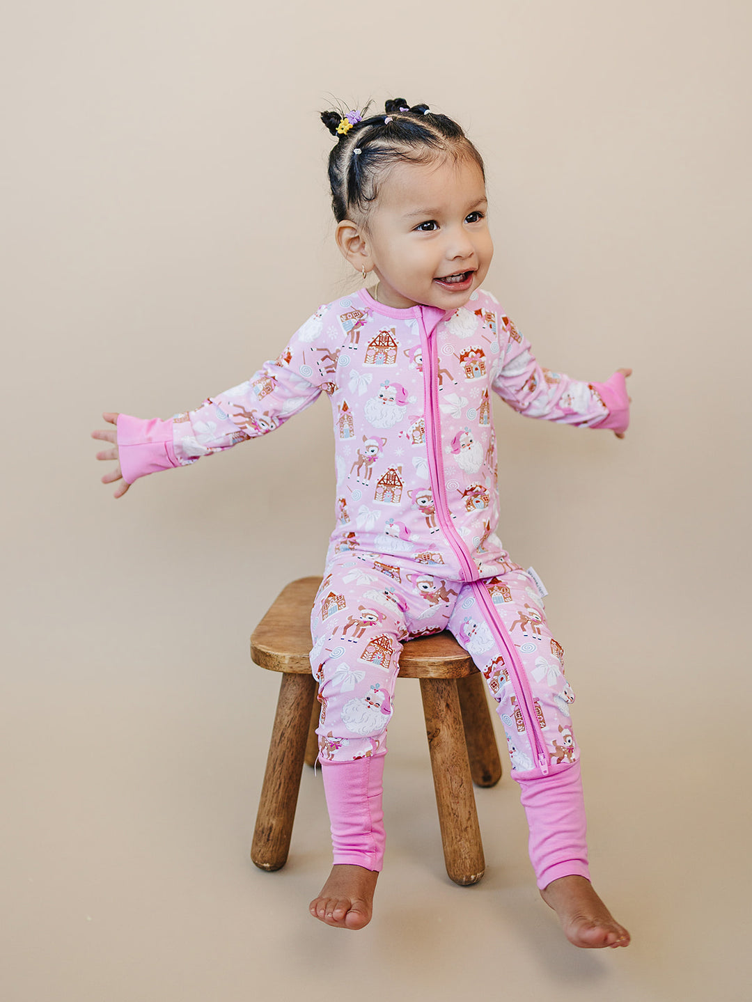Smiling toddler sitting on a wooden stool with arms open in the Pink Christmas zip romper.