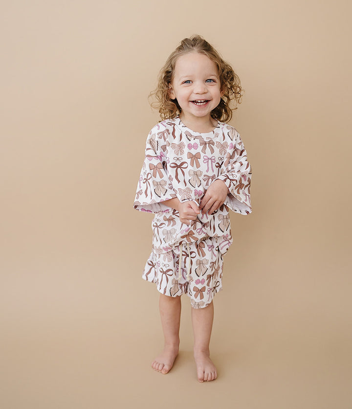 A young girl with curly light brown hair stands barefoot against a solid beige studio background, wearing a matching two-piece pajama set with a white base and a brown bow and ribbon pattern.