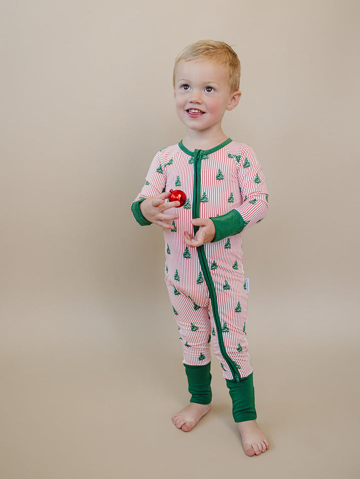 Smiling toddler holding a red ornament in the Magical Trees Christmas zip romper with green ankle cuffs.