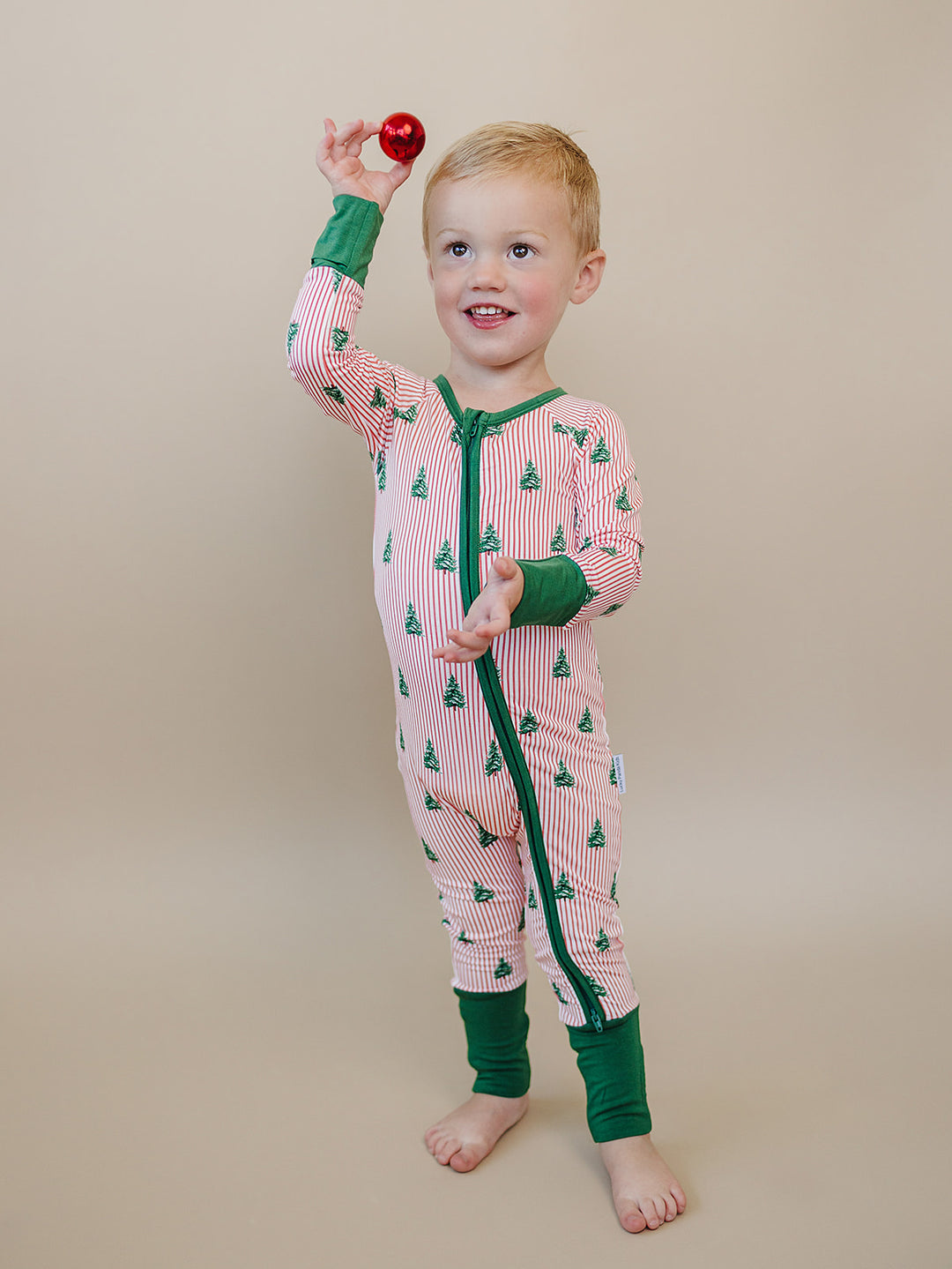 Toddler holding a red ornament overhead while wearing the Magical Trees Christmas romper with green trim.