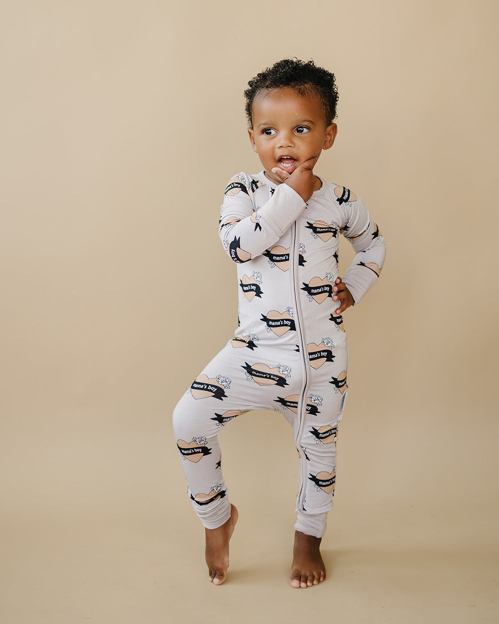Smiling baby lying on a cream quilt in the Mama’s Boy bamboo zip romper with beige hearts and black banners.