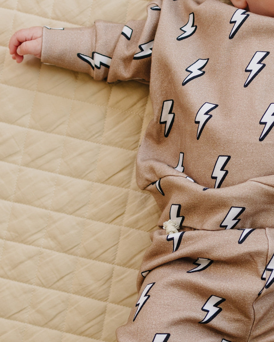 A close-up of a baby wearing a beige long-sleeve romper with a repeating white and black lightning bolt pattern, lying on a quilted beige mattress.