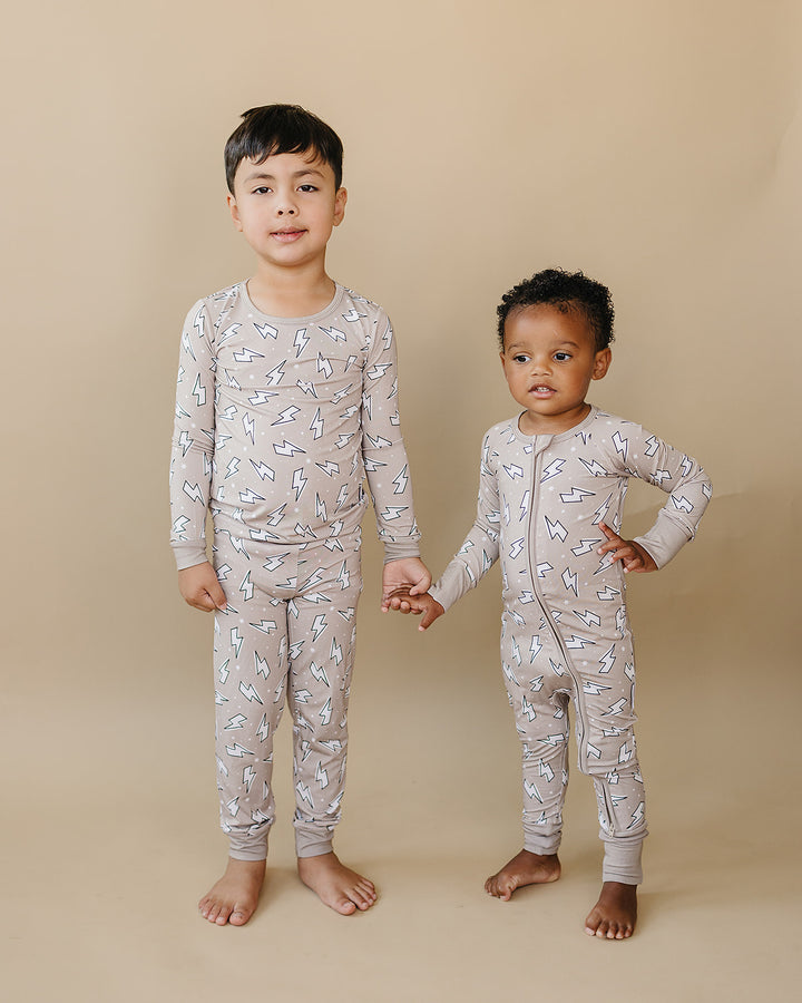 Two boys standing together in matching Latte Bolts bamboo zip rompers against a warm beige studio background.