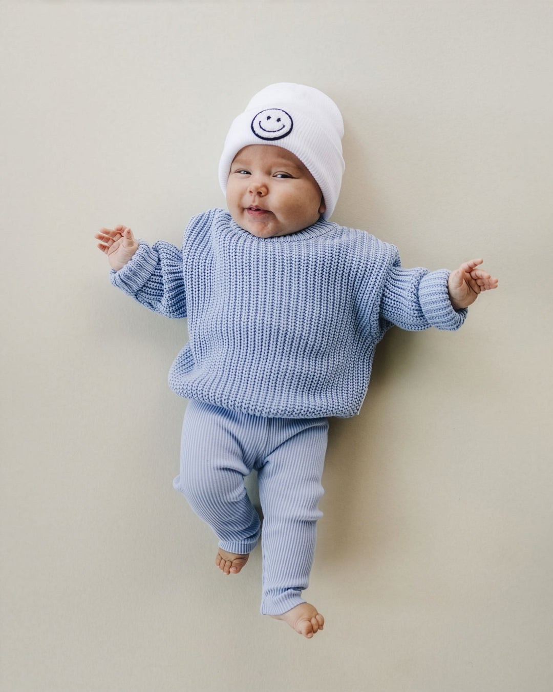 A baby lying on their back wearing a light blue ribbed knit sweater and matching pants, along with a white knit beanie featuring a dark blue smiley face logo, against a solid beige background.