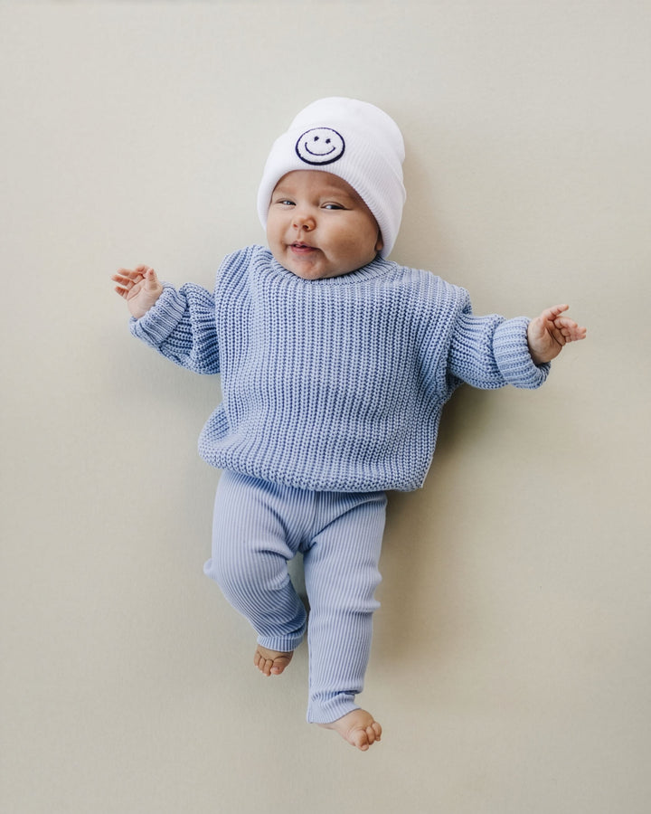 A baby lying on their back wearing a light blue ribbed knit sweater and matching pants, along with a white knit beanie featuring a dark blue smiley face logo, against a solid beige background.
