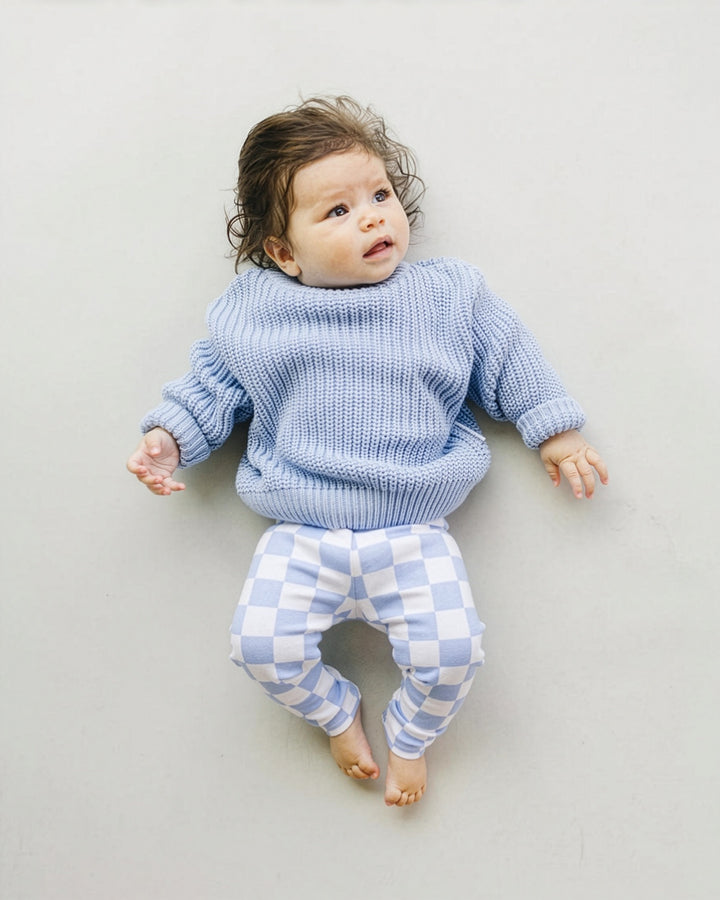 A baby lying on their back wearing a light blue ribbed knit sweater and matching blue and white checkered pants, photographed against a neutral light gray background.