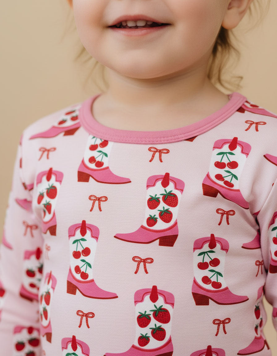 Child wearing pajamas with pink cowboy boots, red cherries, and bows on a white background.