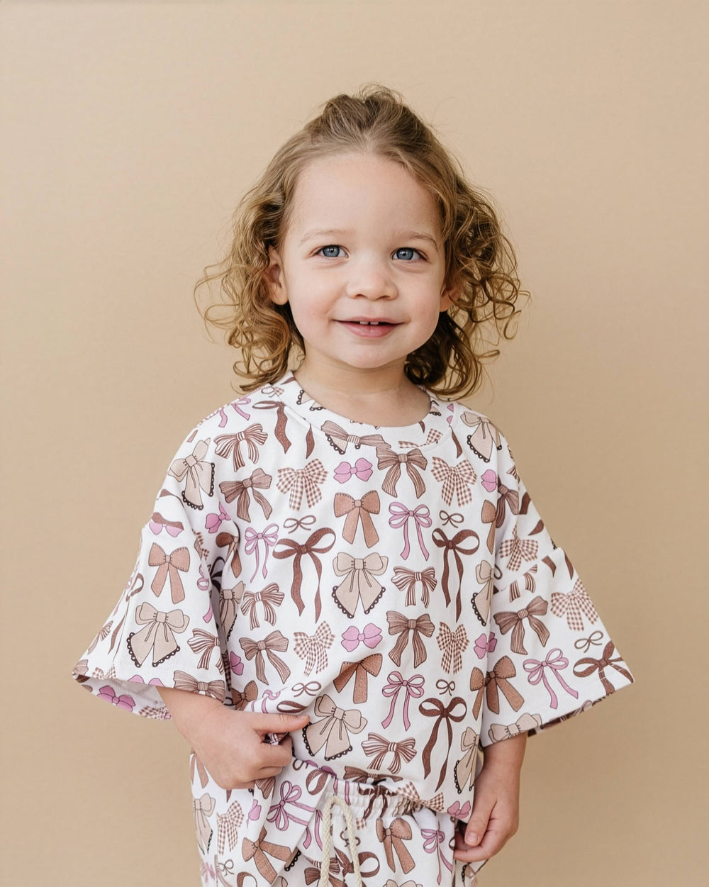 A young child with curly light brown hair wearing a matching two-piece pajama set. The pajamas feature an all-over print of brown and pink bows, ribbons, and hearts on a white background.