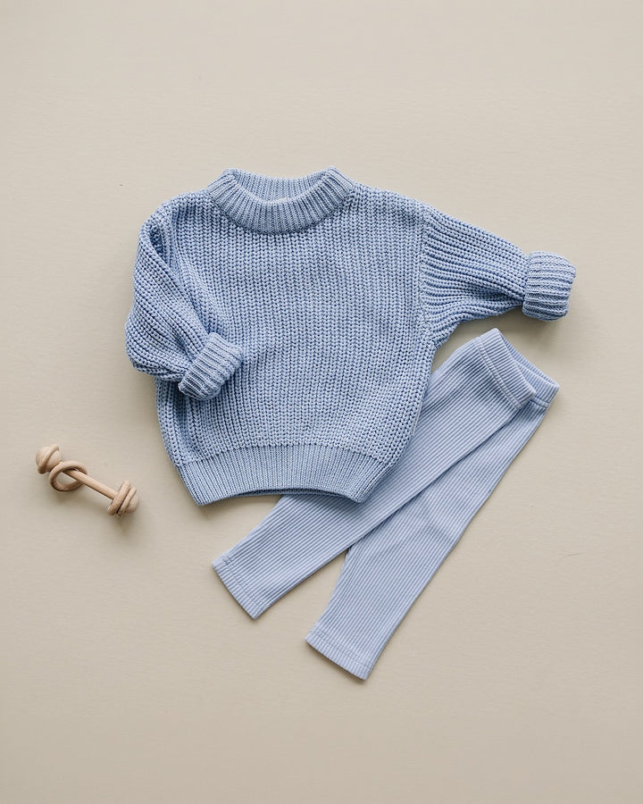 A light blue knit children's sweater and matching leggings set arranged on a warm beige background, with a small wooden toy nearby.