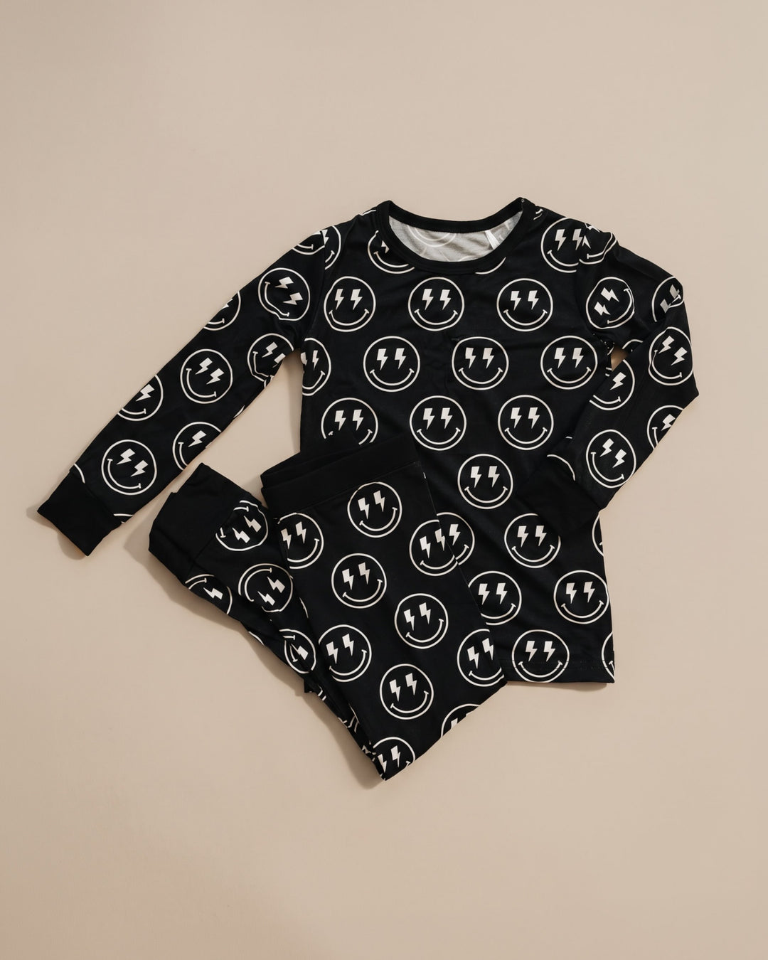 A baby long-sleeve pajama set consisting of a black top and matching pants, both covered in a white circular smiley face pattern with a lightning bolt in the center, laid flat on a beige background.