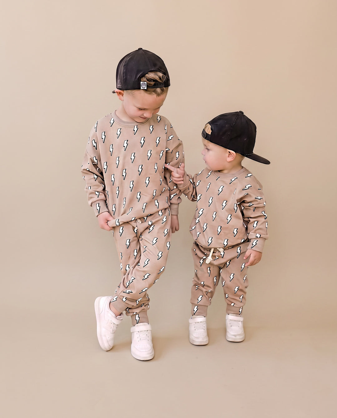 Two toddlers modeling matching beige and brown patterned sweatshirt and jogger sets, wearing black baseball caps and white sneakers, standing against a solid beige studio background.