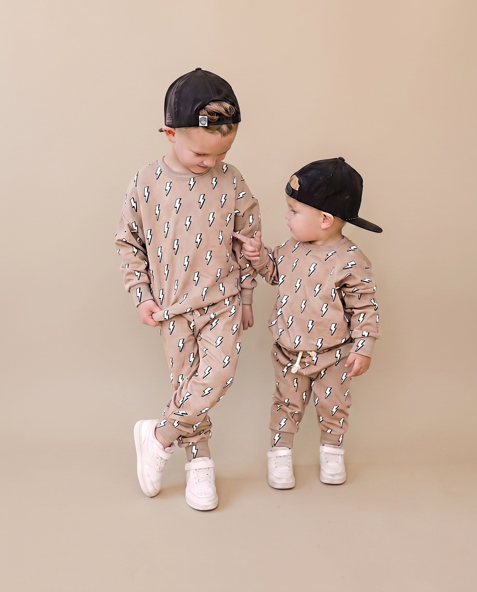 Two toddlers modeling matching beige and brown patterned sweatshirt and jogger sets, wearing black baseball caps and white sneakers, standing against a solid beige studio background.